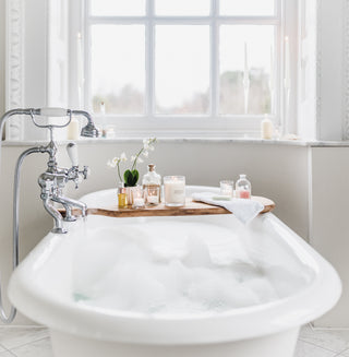 A wooden bath board sits over a relaxing bath with bubbles. On the board is a vase of white orchids, a James and Co Fragrance candle, and a jar of bath salts to make a spa from home.