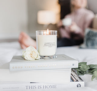 One of our candles sits lit atop a few books. There's a white rose next to the candle, with ambient lighting and a woman drinking from a mug out of focus in the background.