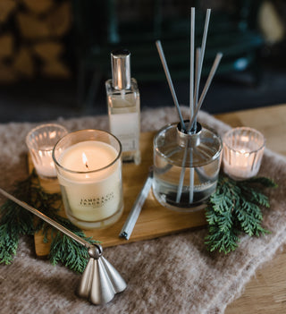 A lit James and Co candle sits on a wooden board, along with a reed diffuser and room spray. There's also a candle snuffer. All of which make great Christmas stocking fillers!
