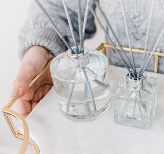 A white womans hand, holding a golden geometric tray, which has two clear glass diffuser bottles with grey reeds sticking out.