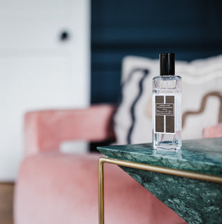 A room spray in a clear bottle with a black label, with a black lid. The product is on a green marble side table. Behind is a chair with a cushion.