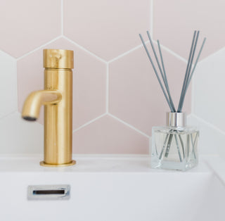 A gold tapped sink with pale pink hexagon tiles behind. To the right is a reed diffuser in a glass jar with grey sticks.