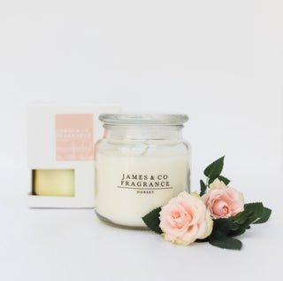 A glass jar candle sitting against a white background, with its box nearby. In front of it are various roses.