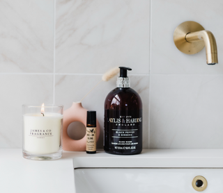 A white jar glass candle which is lit, sitting on the edge of a sink in a bathroom with various bathroom items sitting next to it.