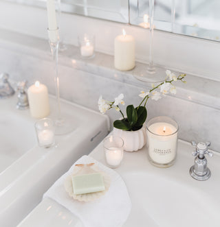 A white jar candle which is lit, sitting on the bathroom sink with various smaller tealights scattered around.