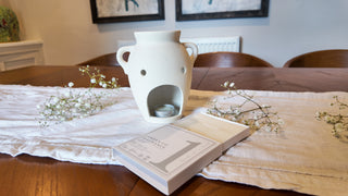Dining table with a white vase, open book, and small plants on a textured tablecloth.