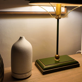 A white, ceramic electric diffuser, sitting on a wooden shelf, next to a lamp.