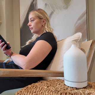 A young, white woman with blonde hair, sitting on her phone next to a white, ceramic electric diffuser.