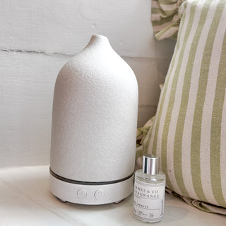 A white electric, ceramic diffuser with a small refill oil sitting next to it on a white shelf.