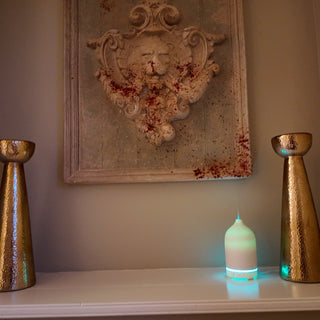 A lit up white ceramic diffuser, sitting on a white shelf, between two gold pillar ornaments.