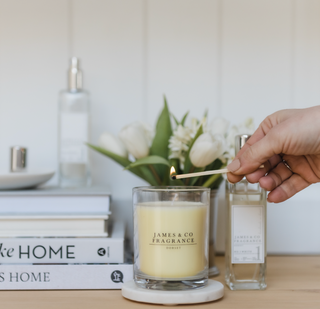 A white female hand, holding a match, lighting a white glass jar candle.