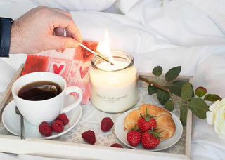 A white male hand, holding a lit match over a tray with various objects including a cup of coffee and a plate with a pastry on it. The hand is lighting a large, white jar candle.