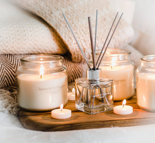 Several lit glass jar candles, with a reed diffuser sitting in between with grey reeds. In the background are cosy blankets.
