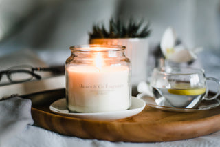A lit glass jar candle sitting on a wooden board with a cup of tea blurred in the background.
