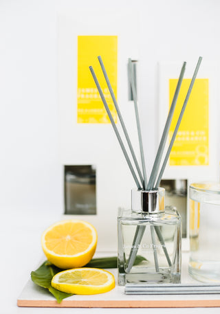 A display of reed diffusers, one out of the box the other two set behind. The packaging is white and yellow. The fragrance is described as lemon. There is a cut fresh lemon to match the scent.