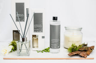 A display of products. two reed diffusers, one small. one larger. A jar candle, unlit, with the glass lid on. There is dried mixed foliage scattered around the image.