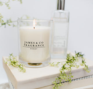 A clear jar candle sitting upon a book. The candle is lit, there is some green foliage scattered around the product.