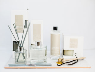 A display of products. two reed diffusers, one small. one larger. A jar candle, unlit, with the glass lid on. There is dried mixed foliage scattered around the image.