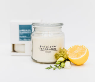 Our white jar glass candle sitting on against a white background, with its box behind it. In front of it sits a slice of lemon.