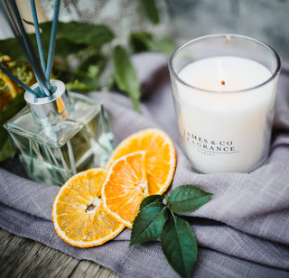 A white glass jar candle, sitting on a grey fabric next to several pieces of orange sliced.