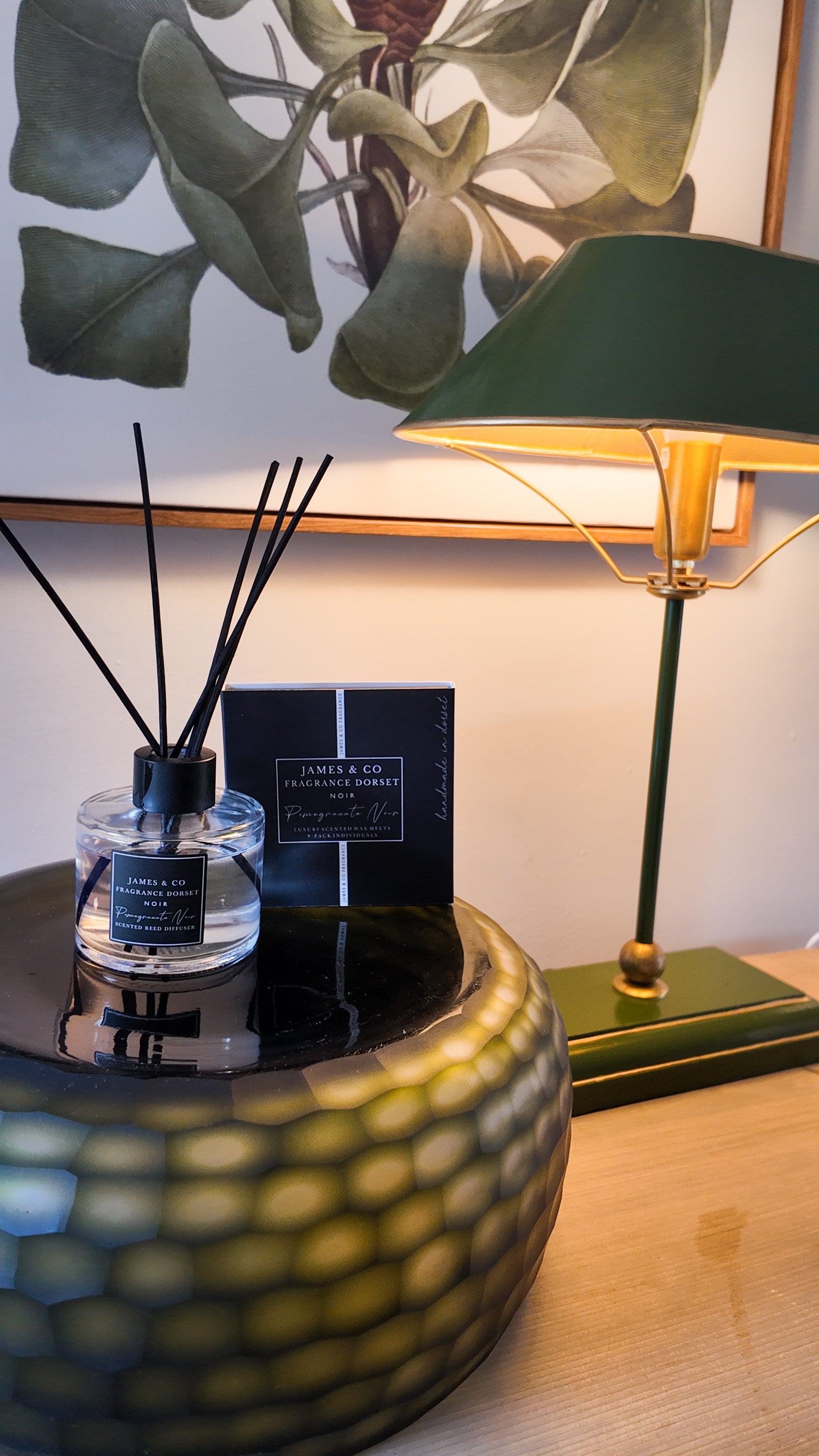A black box, next to a glass diffuser with black accents, sitting on a green bowl on a wooden desk next to a lamp.