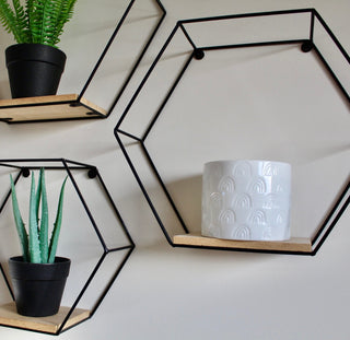 A white ceramic candle with a rainbow pattern.The candle is sitting inside a black hexagon shelf, there are three shelves. The other two have some faux potted greenery.