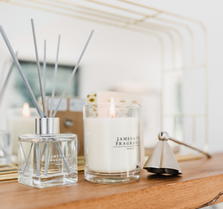 A reed diffuser in a clear bottle with grey sticks sitting on a wooden shelf. Next to the diffuser is a white candle in a glass jar, the candle is lit. There is a candle snuffer in chrome.