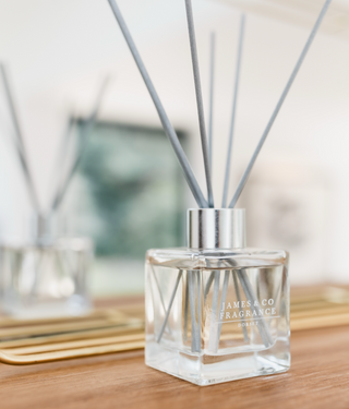 A square reed diffuser in a glass jar with grey sticks. The diffuser is sitting on a wooden table and a mirror is directly behind.