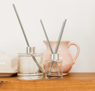 Two reed diffusers, one small and square, the second larger and round. Both have grey sticks. The products are sitting on a wooden table. behind them is a pink ceramic jar.