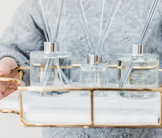 A trio of clear glass reed diffusers, two large one small in the middle. All have grey sticks. They are sat on a gold and white tray with a lady in a grey jumper displaying them.