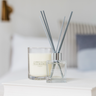 A white candle in a glass jar, the candle is un lit. Just in front sits a reed diffuser also in a glass jar with grey sticks. The objects are both being displayed on a white bedside table.