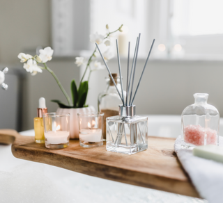 A wooden bath tray displaying a clear glass reed diffuser, two small lit candles. A glass small jar containing pink bath salts. There is some face oil and a artificial plant in a ceramic vase.