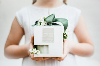 A child wearing a white dress, holding the boxed No.7 Vanilla jar candle with a green bow and white flowers around it.