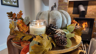 A white candle, in a glass jar, sitting on a gold table with leaves and foliage scattered around it. In the background is a roaring log fire.