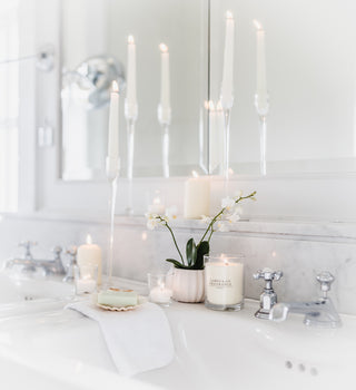 A white glass jar candle which is lit, sitting on a bathroom sink