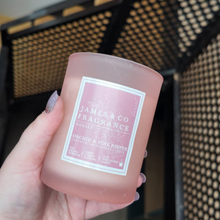 A pink candle, in a frosted glass jar with a pink label, being held against a blurred background.