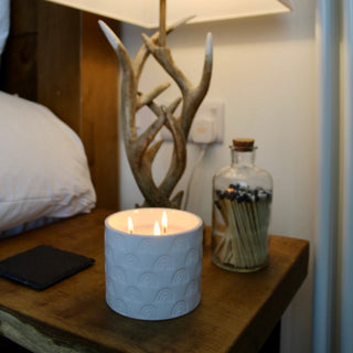 A ceramic rainbow candle on a wooden table. The candle is lit and has three wicks. Behind is a glass jar with matches inside. There is lamp on the table that is tuned on.