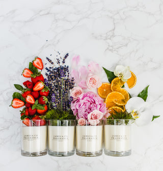 Four James & Co Fragrance candleslaid down on a marble surface with various garnishes above them. From left to right: 1 has Strawberries; 2 has lavender; 3 has pink roses; 4 has slices of oranges and white orchids.