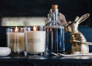 Two lit candles with 'James & Co Fragrance' branding, a bottle of perfume, and a gold rabbit figurine on a dark surface.