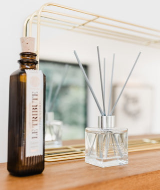 A bottle and a diffuser on a wooden surface infront of a gold frames mirror.