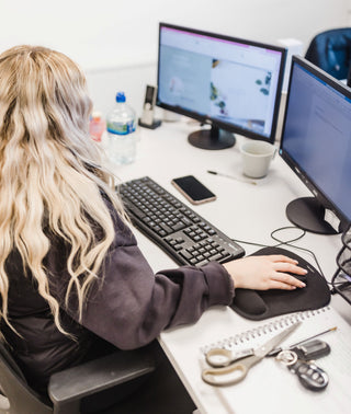 Person sitting at a desk with two computer monitors, keyboard, and various items on a white surface.
