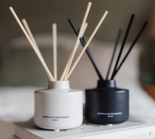 Two diffusers with sticks on a book, one white and one black, on a soft surface.