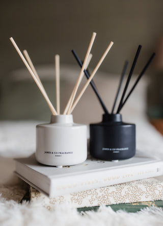 Two diffusers with sticks on a book, one white and one black, on a soft surface.
