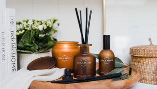 Set of home fragrance products including diffusers and candles on a wooden surface with a book and flowers in the background.