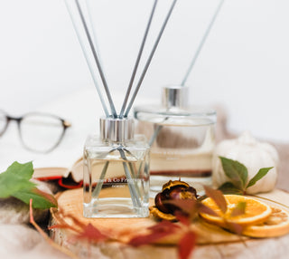 Glass diffuser bottle with grey reeds, sat on a wooden tray with autumn leaves and orange slices
