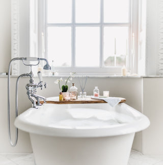 White freestanding bathtub with a wooden tray holding bath products in a bright bathroom.