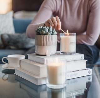 Person lighting a candle on a stack of books with a small potted plant nearby.