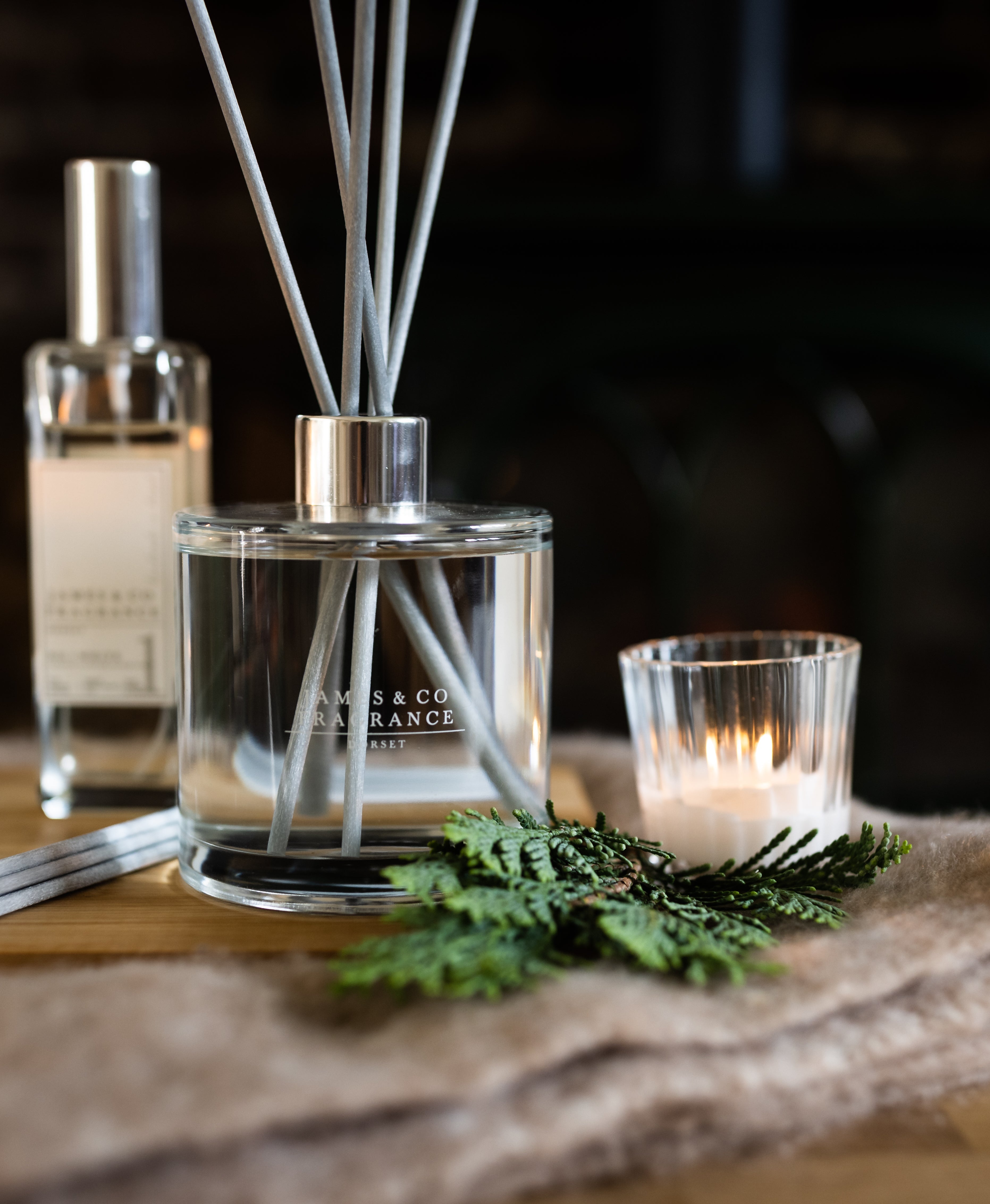 A large round reed diffuser sits next to a tealight candle on green leaves and a dimly lit room