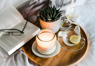 A lit white glass jar candle sitting on a wooden tray with a book, a plant and a cup of tea nearby.