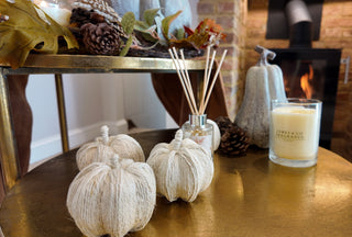A gold table, with a lit fire in a log burner in the background. On the table sits various pumpkins which are scented, along with a candle and seasonal accessories. 